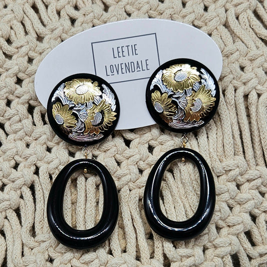 Pair of earrings with black and gold design on a textured beige surface, branded 'LEETIE LOVENDALE'.