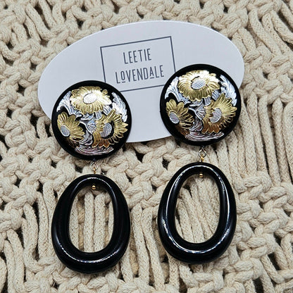Pair of earrings with black and gold design on a textured beige surface, branded 'LEETIE LOVENDALE'.