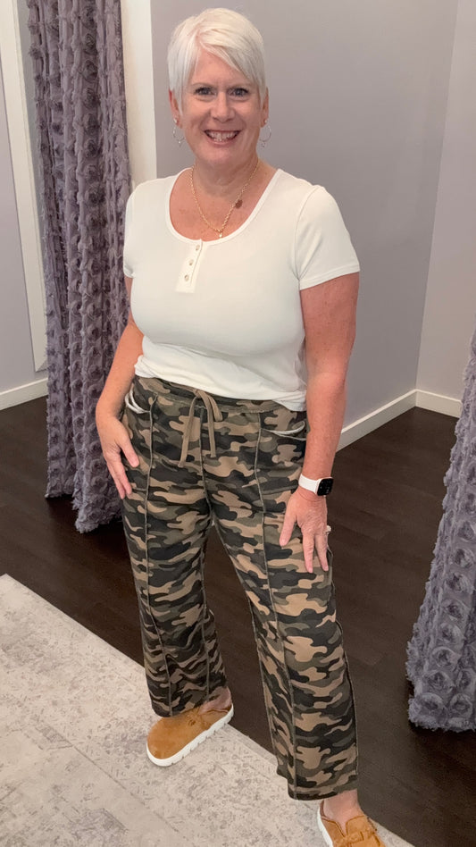 A woman with short white hair stands indoors wearing a white short-sleeve henley top and camouflage drawstring pants, paired with tan slip-on shoes. She is posed in a room with gray walls, patterned curtains, dark wood flooring, and a light rug.