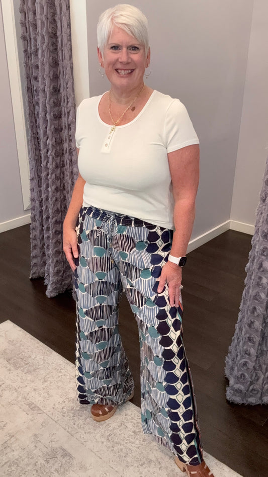 A woman with short white hair stands in a fitting room wearing a white short-sleeve henley top and high-waisted, wide-leg trousers with a blue, gray, and white abstract pattern.