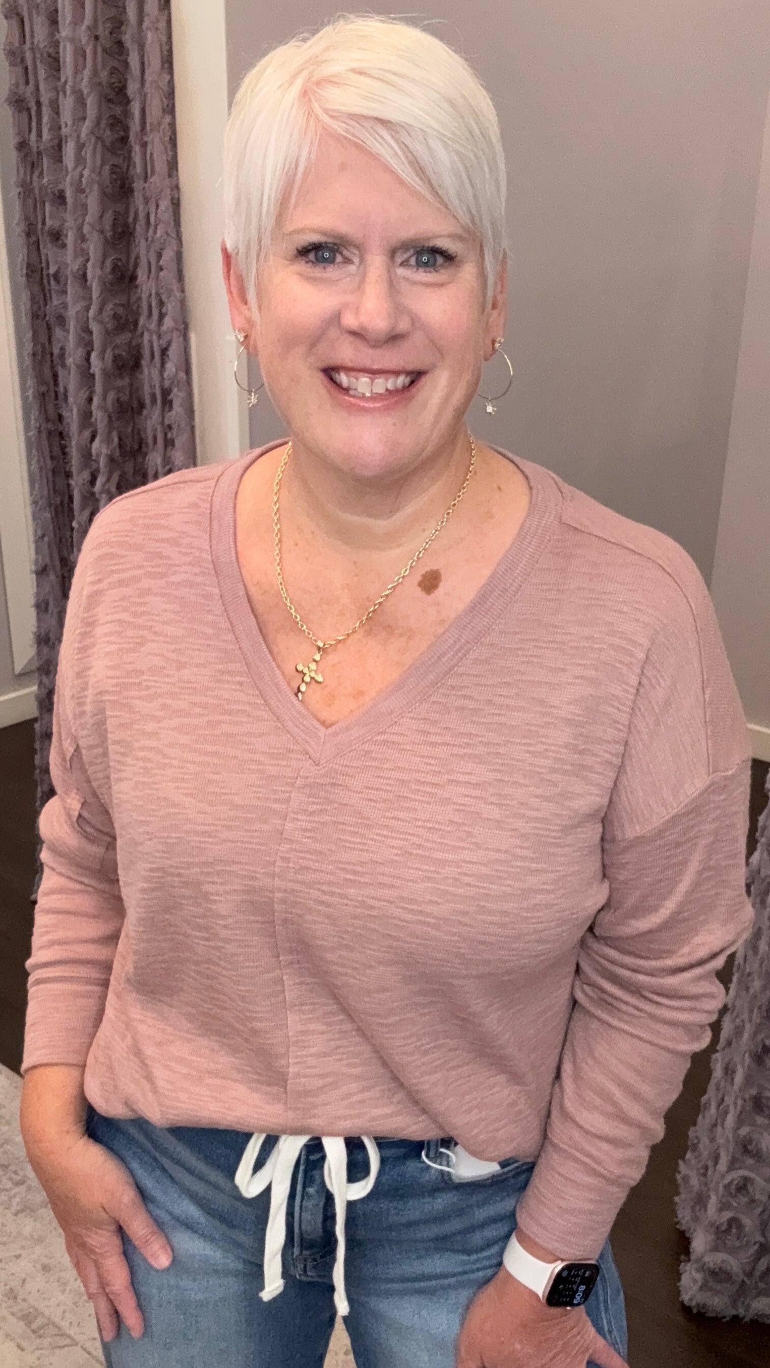 A smiling woman with short platinum blonde hair wearing a dusty rose pink long-sleeve V-neck top and blue jeans with a white drawstring, standing indoors.