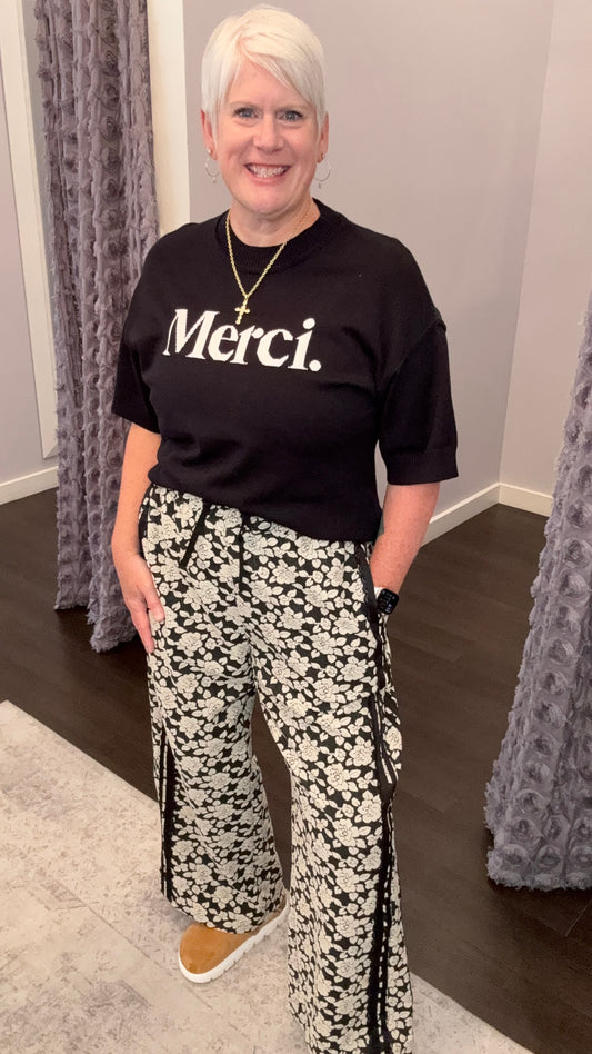 A woman with short white hair stands in a fitting room wearing a black t-shirt with 'Merci.' printed in white across the chest, paired with black and white floral wide-leg pants and tan slip-on shoes.