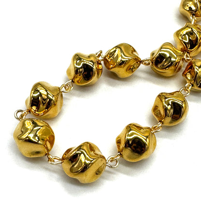 A close-up of a gold-toned beaded necklace with irregular, organic-shaped beads connected by small links, photographed on a plain white background.