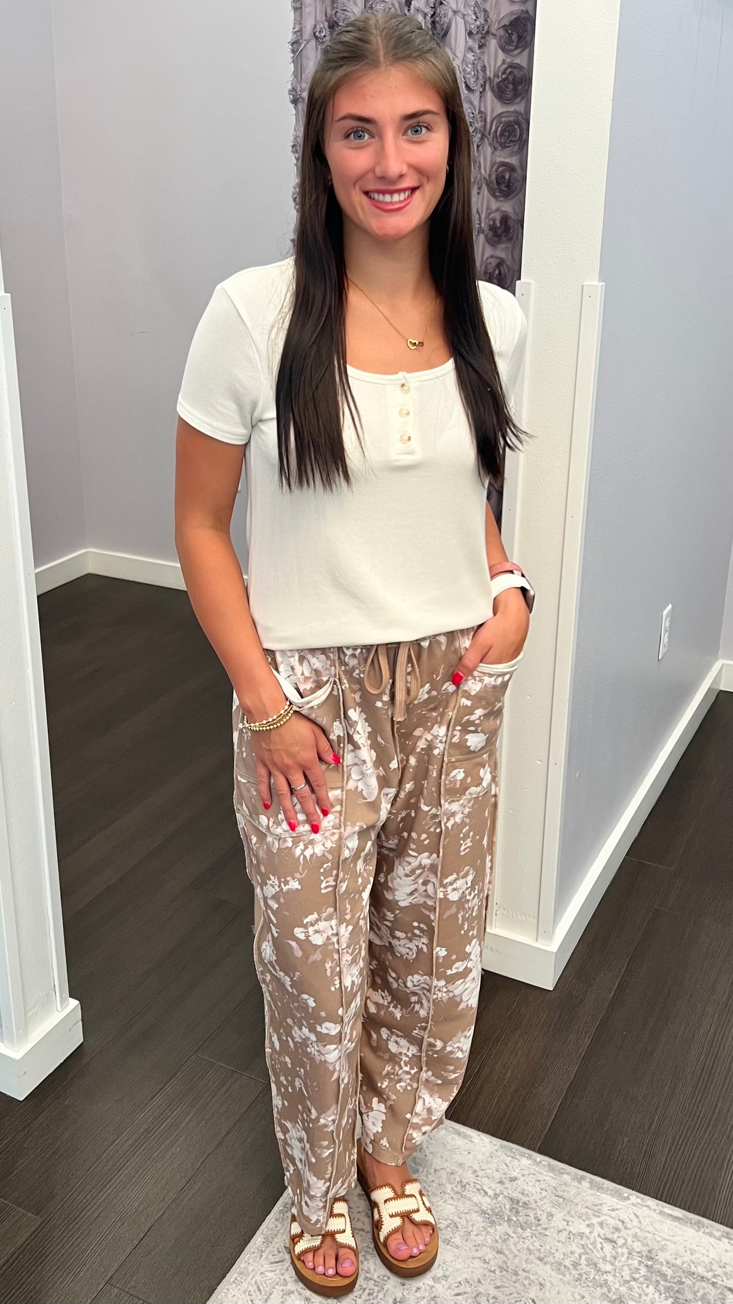 A woman standing indoors wearing a white short-sleeve henley top and beige patterned drawstring pants, paired with tan sandals.