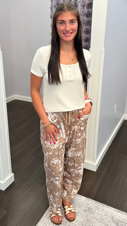 A woman standing indoors wearing a white short-sleeve henley top and beige patterned drawstring pants, paired with tan sandals.